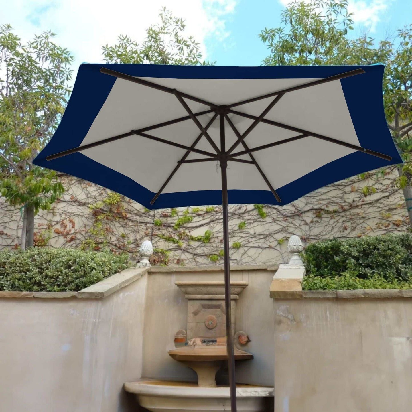 A 9ft Market Patio Umbrella featuring the 6 Rib Replacement Canopy Duet Navy from Formosa Covers is open, providing shade over a stone fountain. The background showcases lush greenery and a stone wall adorned with climbing plants. The sky is partially visible, indicating a clear day.