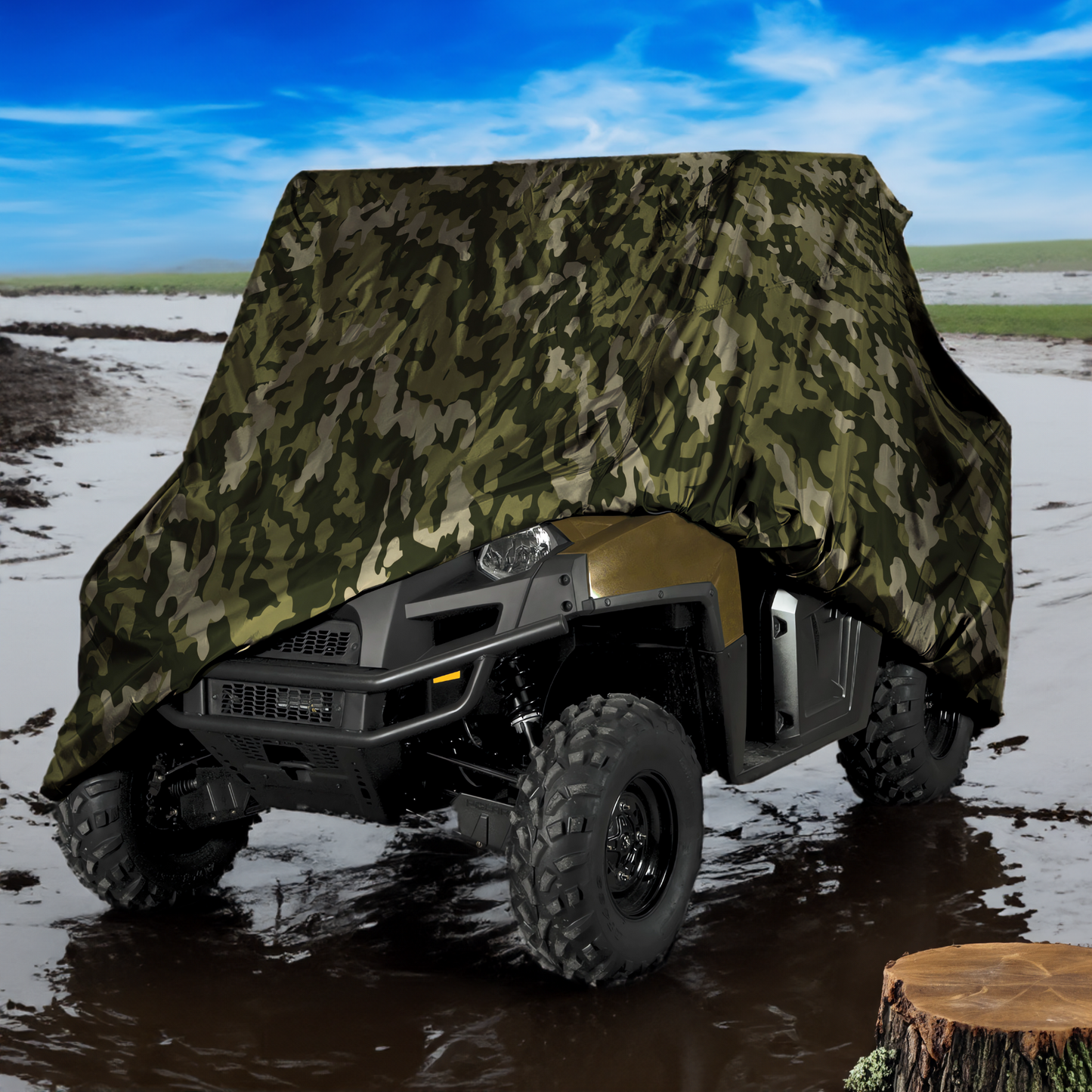 A rugged all-terrain vehicle with large off-road tires is partially covered by a heavy-duty, UV-treated, water-repellent camouflage tarp from Formosa Covers. The UTV Cover - Large Camouflage 120" L x 62" W x 75"H blends seamlessly into the surroundings as it sits parked on muddy ground, with a bright blue sky and open landscape in the background, suggesting a remote or wilderness setting.
