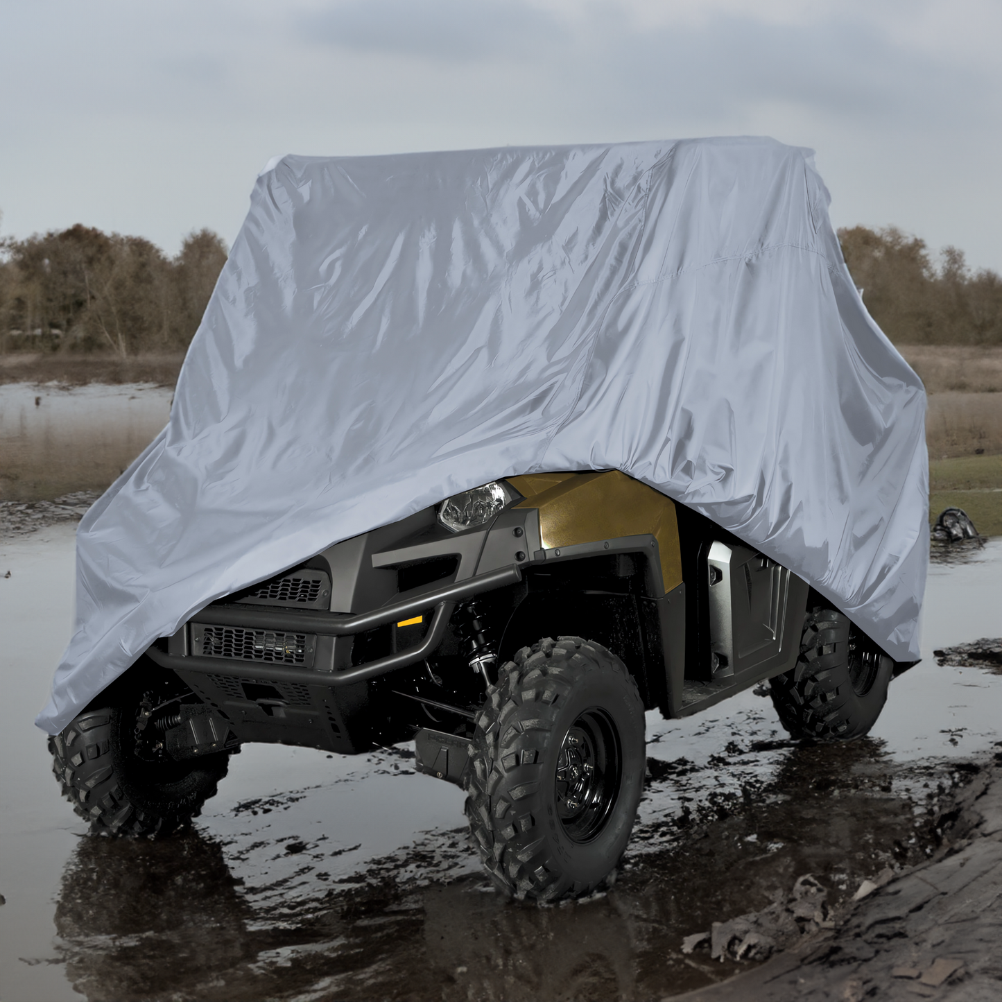 A rugged, off-road vehicle is partially covered with a heavy-duty, gray UTV Cover - Large 120" L x 62" W x 75"H from Formosa Covers. The vehicle is positioned in a muddy, rural area with bare trees in the background under an overcast sky.