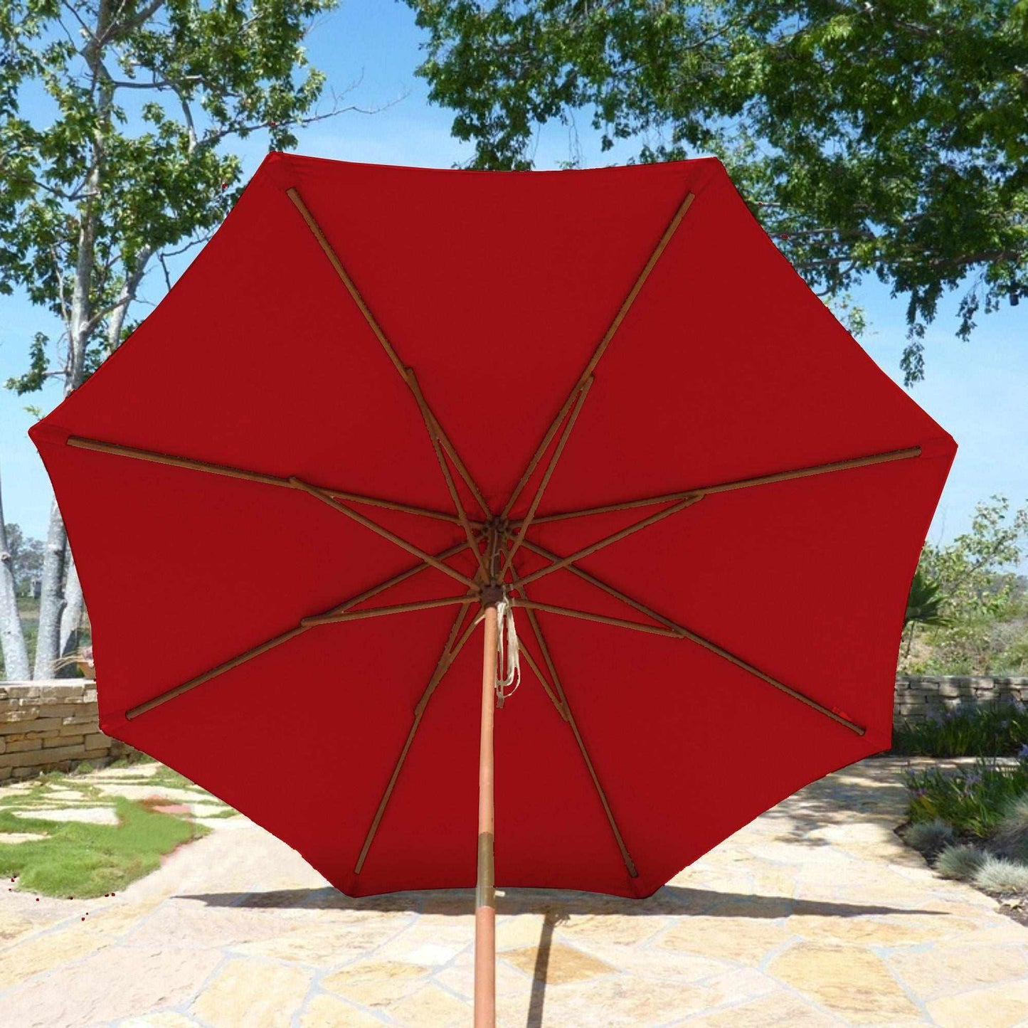The 9ft Market Patio Umbrella 8 Rib Replacement Canopy in Cherry Red by Formosa Covers is open on a sunlit stone patio, showcasing its UV-treated 300 denier polyester fabric. The canopy is supported by wooden ribs, and the base is set centrally on the patio. Green trees and foliage provide a lush background.