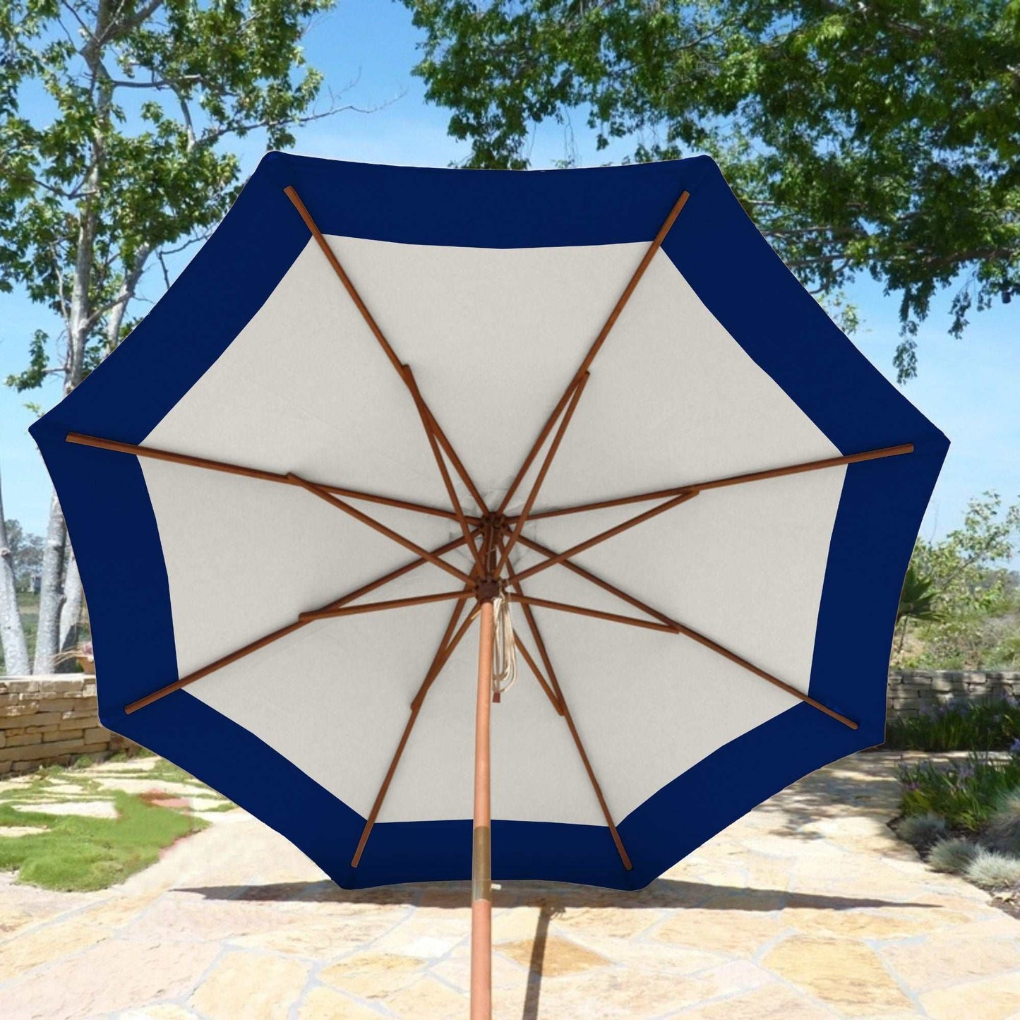 The 9ft Market Patio Umbrella 8 Rib Replacement Canopy Duet Navy by Formosa Covers is open, providing shade with its UV-treated canopy. It stands on a wooden pole and is set on a stone-paved patio with green trees and plants in the background. The umbrella features a sturdy wooden frame supporting its fabric.
