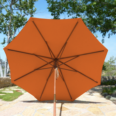 The 9ft Market Patio Umbrella 8 Rib Replacement Canopy in Tuscan Orange, from Formosa Covers, is open, providing shade on a sunny day. The vibrant umbrella stands on a stone-paved area with greenery and trees in the background. The sky is clear and blue.