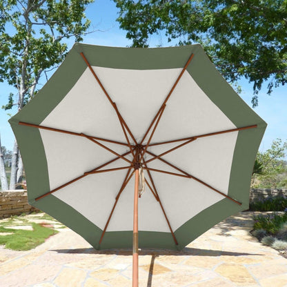 A 9ft Market Patio Umbrella from Formosa Covers, featuring a sage green border and white center made from durable polyester fabric, is opened on a stone patio. The umbrella boasts a wooden frame and pole. In the background, there are trees and a clear sky.