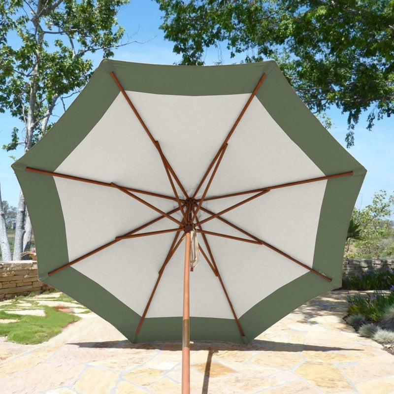 A 9ft Market Patio Umbrella from Formosa Covers, featuring a sage green border and white center made from durable polyester fabric, is opened on a stone patio. The umbrella boasts a wooden frame and pole. In the background, there are trees and a clear sky.