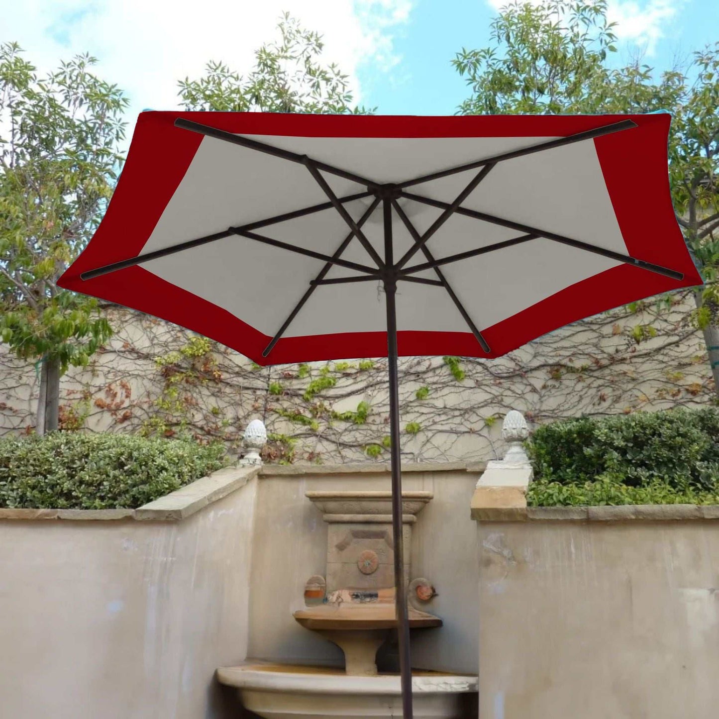 A 9ft Market Patio Umbrella 6 Rib Replacement Canopy Duet Red by Formosa Covers, made from 300 denier polyester fabric, is open, providing shade over a garden area with a stone water fountain. The backdrop features a light-colored, vine-covered wall and lush greenery, with a partly cloudy sky above.