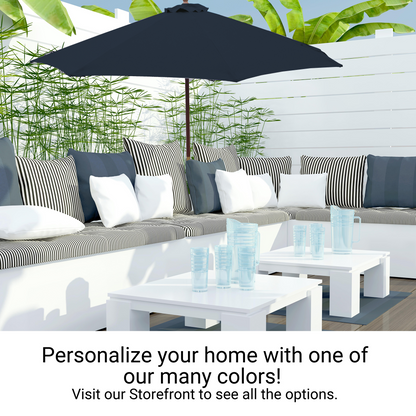 A modern outdoor patio with white furniture, including a sectional sofa with striped and solid cushions, white tables, and scattered transparent cups. A large dark blue 9ft market patio umbrella from Formosa Covers provides shade with its 8 rib canopy, and lush green plants create a serene backdrop.