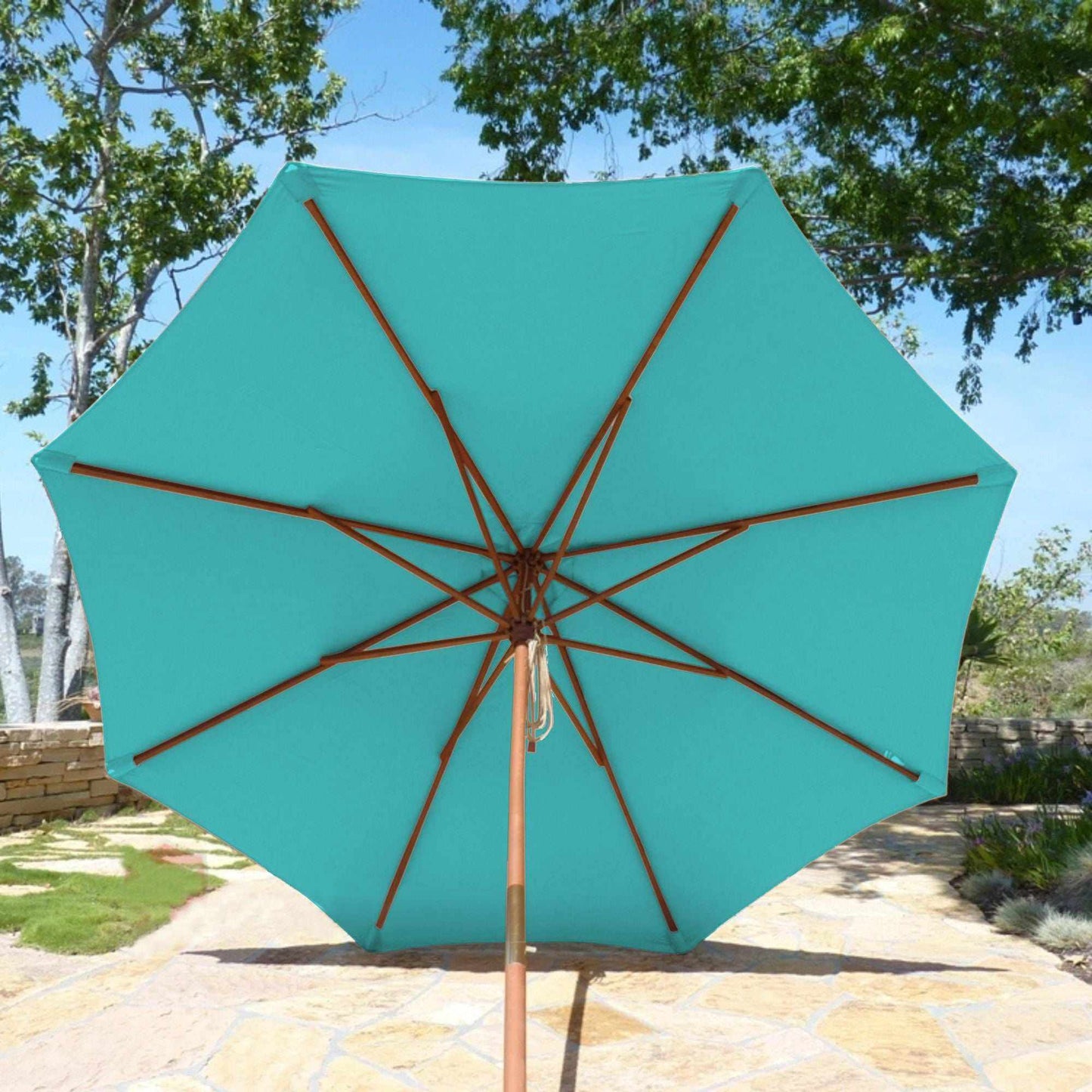 An 11ft Market Patio Umbrella, featuring an Aruba Turquoise canopy with 8 ribs from Formosa Covers, is set up on a stone patio in a garden with lush greenery and trees in the background on a sunny day. The umbrella boasts durable polyester fabric, UV-treated for long-lasting protection, and wooden support poles.