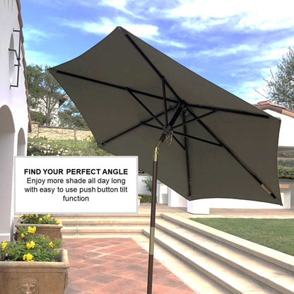 A 7ft Wooden Patio Garden Market Umbrella with Tilt Mechanism by Formosa Covers, featuring a sage green canopy and wooden crank and tilt, is angled to the right, providing shade on a sunny day with a partly cloudy sky. A sign next to the umbrella reads, "FIND YOUR PERFECT ANGLE - Enjoy more shade all day long with an easy-to-use push button tilt function.