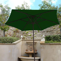 A 9ft Market Patio Umbrella with a black frame and Forest Green UV-treated durable polyester fabric canopy from Formosa Covers stands open in an outdoor garden area. The background features a stone wall adorned with climbing plants, green foliage, and a stone water fountain. The sky above is clear with a few clouds.