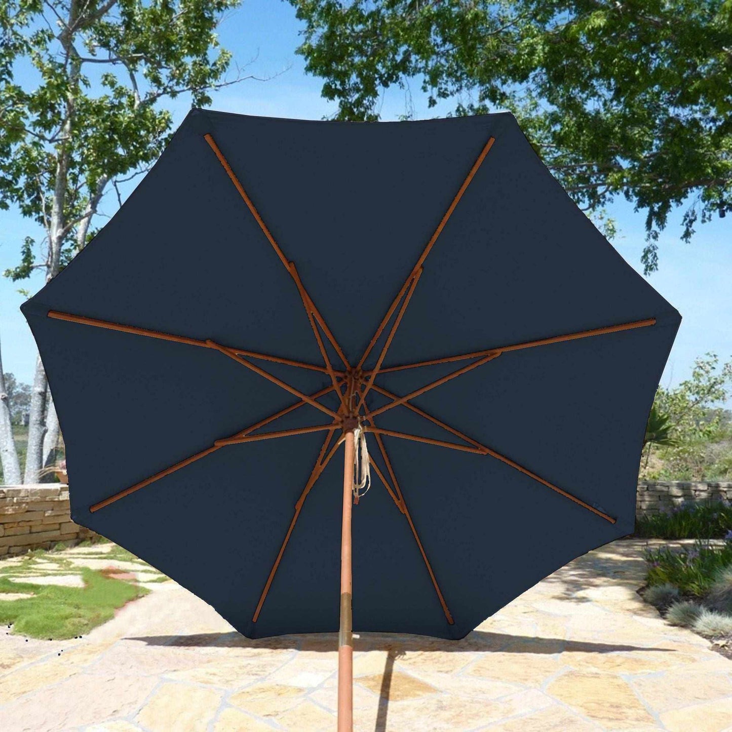 An 11ft Market Patio Umbrella with an 8 Rib Replacement Canopy in dark blue from Formosa Covers is open and providing shade. The durable patio umbrella features a wooden pole and spokes and is set on a stone-paved patio, surrounded by lush green trees and a garden. The sky above is clear.