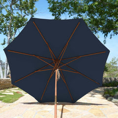 An 11ft Market Patio Umbrella with an 8 Rib Replacement Canopy in dark blue from Formosa Covers is open and providing shade. The durable patio umbrella features a wooden pole and spokes and is set on a stone-paved patio, surrounded by lush green trees and a garden. The sky above is clear.