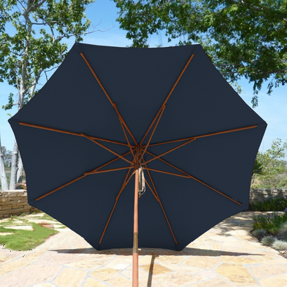 A Formosa Covers 9ft Market Patio Umbrella with an 8 rib replacement canopy in dark blue is open on a sunny day, casting shade over a paved outdoor area. The wooden frame of the UV-treated umbrella is visible, and trees with green foliage surround the space, creating a serene and inviting atmosphere.
