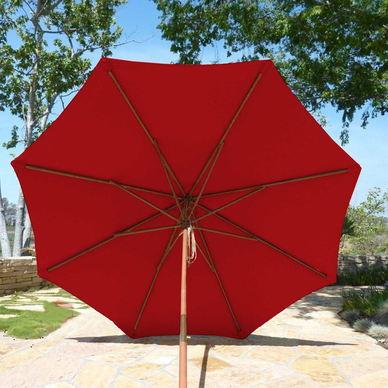 The 9ft Market Patio Umbrella 8 Rib Replacement Canopy in Cherry Red by Formosa Covers is open on a sunlit stone patio, showcasing its UV-treated 300 denier polyester fabric. The canopy is supported by wooden ribs, and the base is set centrally on the patio. Green trees and foliage provide a lush background.