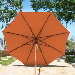 A large, tangerine 9ft market patio umbrella with a durable polyester fabric canopy by Formosa Covers is seen from below on a bright day. The scene features a stone-paved outdoor area surrounded by greenery, including trees and shrubs, adding to the natural beauty of the setting.