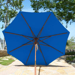 A 9ft Market Patio Umbrella with an 8 Rib Replacement Canopy in Cobalt Blue from Formosa Covers is open, casting shade over a stone patio. Set against a backdrop of lush greenery, including trees and bushes, this stylish umbrella features eight spokes supporting its structure and is equipped with a wooden pole.