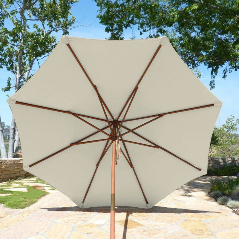 A 9ft Market Patio Umbrella with an 8 Rib Replacement Canopy in Natural by Formosa Covers is open on a sunny day, casting shade on a stone pathway. The umbrella features a wooden pole and rib structure, set against a backdrop of green trees, a lawn, and a stone wall.