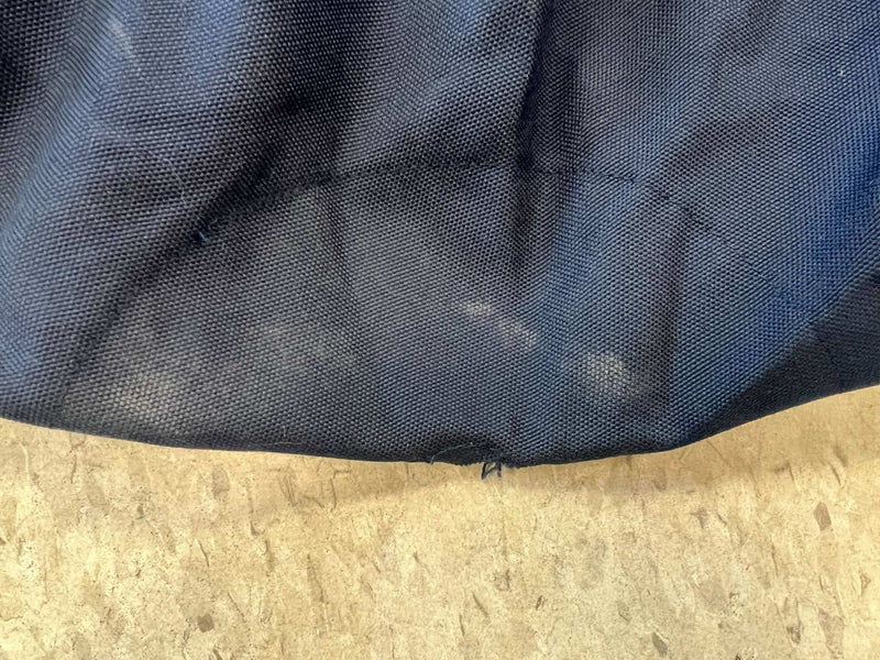 A close-up of a small hole or tear in the navy fabric made from Premium Olefin material of the 9ft Market Patio Umbrella 8 Rib Replacement Canopy by Formosa Covers. The damage is located near what seems to be the edge or seam. The background is a light-colored, textured surface, possibly a floor or tabletop, highlighting the UV-treated and water-repellent properties.