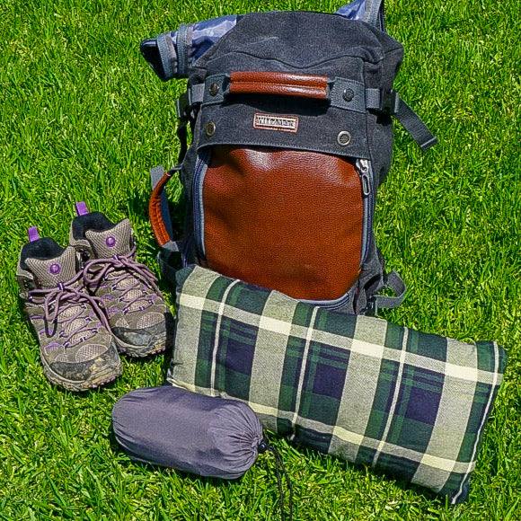A neatly arranged outdoor gear set lies on green grass: a grey and brown hiking backpack, a rolled-up green and white plaid blanket, a pair of worn hiking boots with purple laces, a Formosa Covers Rectangular Sleeping Bag Liner for Hiking, Camping, or Hostel use with an 80" zippered length, and a compact rolled-up grey sleeping bag.