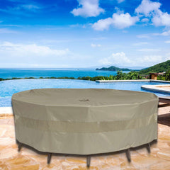 The stone patio, offering a scenic view of an infinity pool, lush green hills, and a distant ocean under a blue sky with fluffy clouds, is adorned with the Classic Taupe Patio Set Cover for Round or Square Table & Chairs by Formosa Covers, providing protection with its UV-treated polyester fabric.