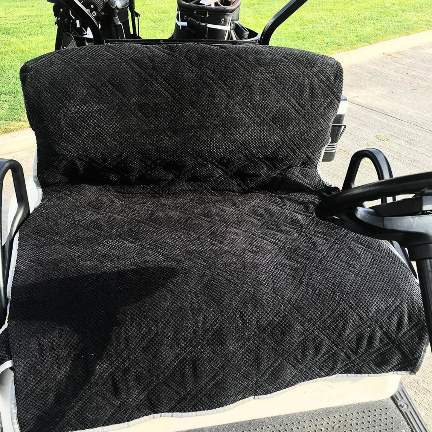 A Reversible Golf Cart Dual Seat Blanket Cover in Black and Taupe by Formosa Covers is draped over the seat. The steering wheel is visible to the right, and part of the grassy area surrounding the cart is seen in the background.