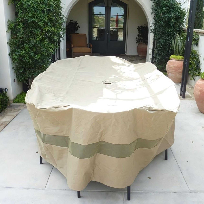 A classic taupe Patio Set Cover for an oval or rectangular table, measuring 110"L x 65"W x 38"H and made by Formosa Covers, is draped over a round table to protect it from the elements. The table is placed on a patio adorned with potted plants and an archway that leads to a door in the background. Under the archway, a cushioned chair can be seen.