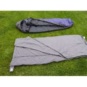 Two sleeping bags are laid out on a grassy lawn. The top one is dark-colored with a hood, partially unzipped to show its interior. The bottom one, the Formosa Covers Sleeping Bag Liner Hiking Camping Hostel Travel Sack Sheet (Rectangular with Zipper, 80"L), is lighter-colored, fully unzipped and spread open, revealing a travel sleep sheet inside.