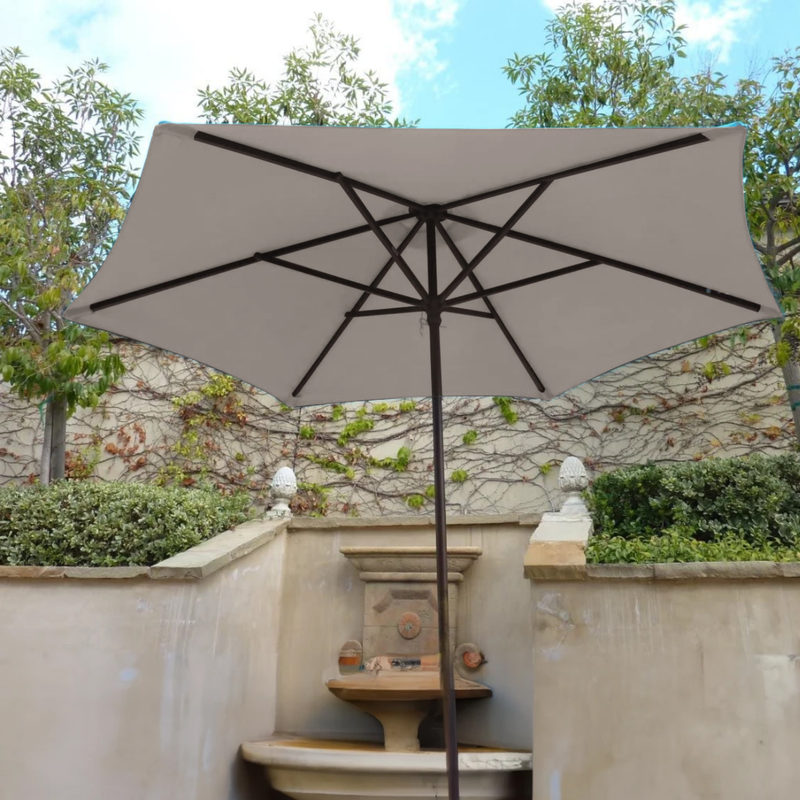 A large 9ft Market Patio Umbrella with a Sand-colored 6 rib canopy from Formosa Covers stands open in a garden area. The umbrella is set against a backdrop of green shrubs, climbing ivy, and a stone water fountain with a rustic wall in the background. Made from durable polyester fabric, it blends perfectly with the partly cloudy sky.